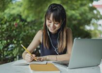 Positive young Asian female student with earphones writing in copybook while doing homework at table with laptop in street cafeteria