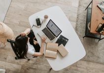 From above of young woman with long dark hair in casual clothes working at table and browsing netbook while sitting in modern workplace and touching hair