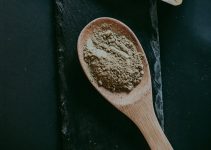 Kratom Maeng Da powder displayed with packet and wooden spoon on a dark surface.