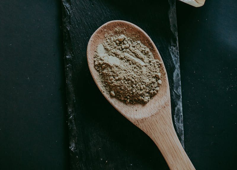 Kratom Maeng Da powder displayed with packet and wooden spoon on a dark surface.