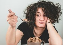 woman with messy hair wearing black crew-neck t-shirt holding spoon with cereals on top
