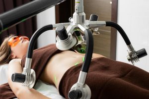 Female client lying on table under lasers of modern weight loss machine on belly during liposuction procedure in medical clinic