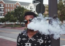 A fashionable man vaping on a street in Williamsburg, VA, with urban scenery.