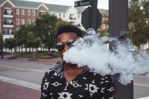A fashionable man vaping on a street in Williamsburg, VA, with urban scenery.