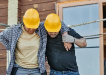 Two construction workers in hard hats assisting each other at a building site, highlighting teamwork.
