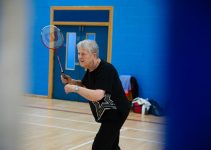 Elderly woman playing badminton in a gymnasium.